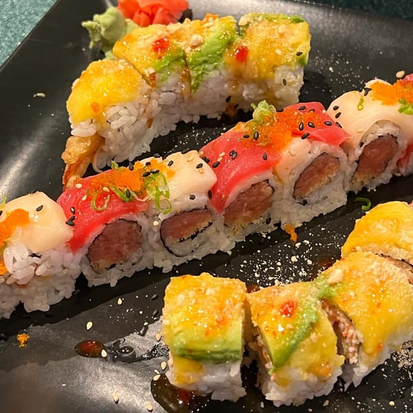 A black plate holds two rows of colorful sushi rolls, garnished with sauces and sesame seeds.